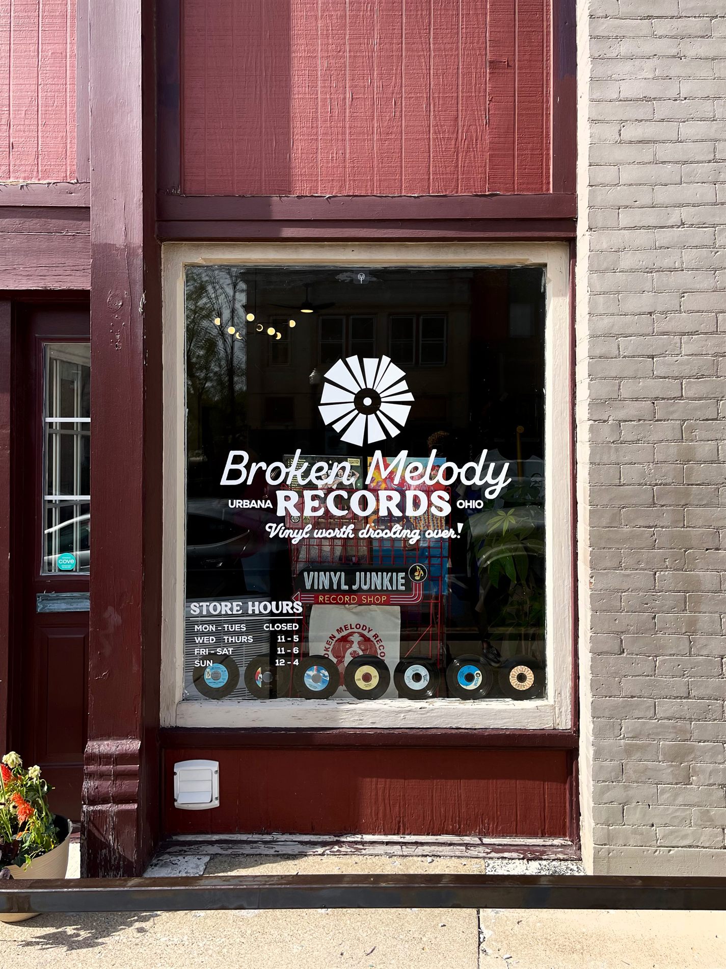 The Broken Melody Records storefront window in Urbana, Ohio, with the logo, tagline, and store hours in white vinyl and a row of 45s along the sill