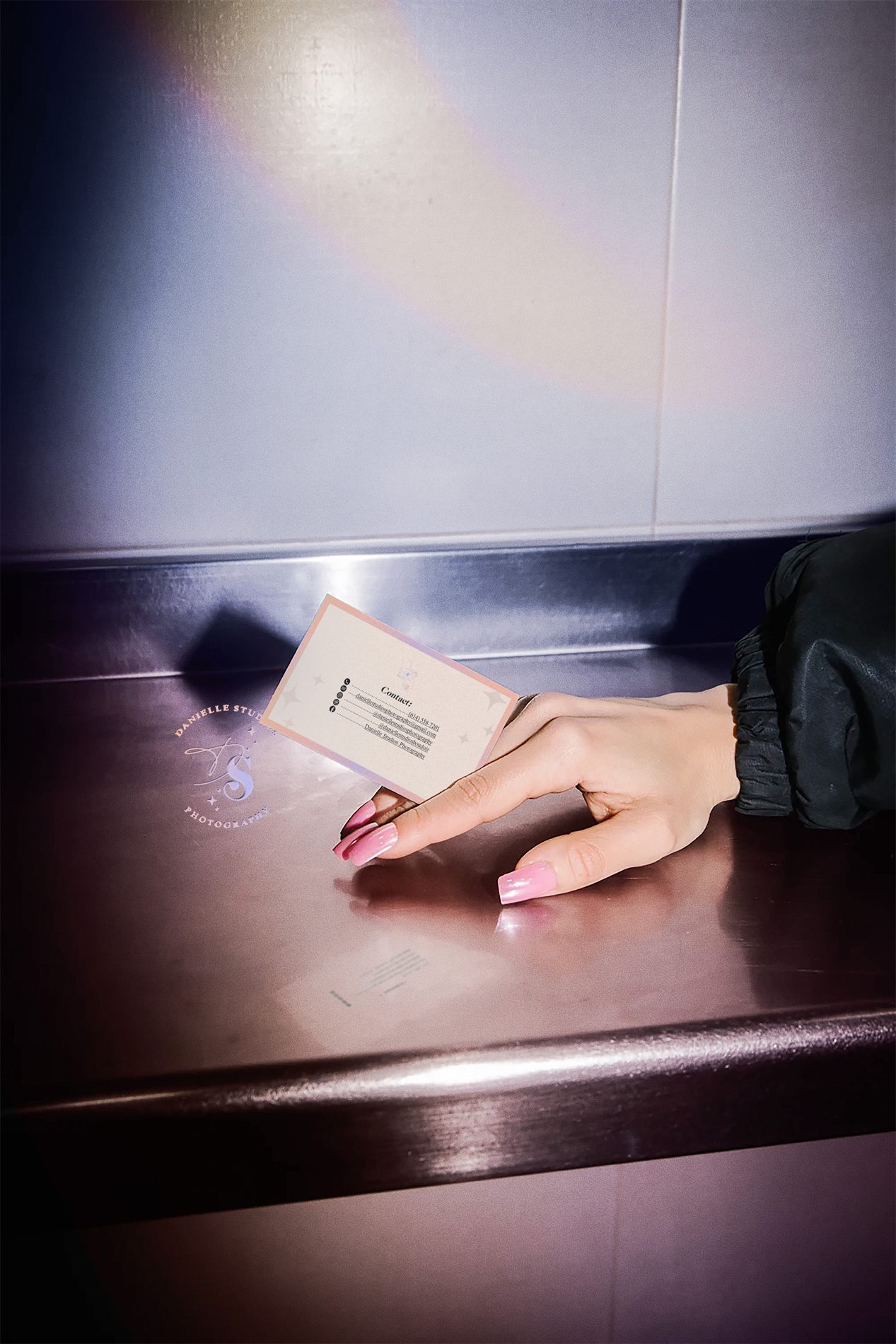 A hand with pink nails holding the Danielle Studios business card, photographed with flash on a metal counter